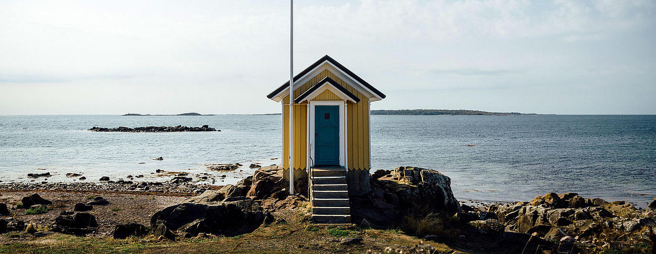 Eine gelb blaue Hütte vor dem Meer. An einem Fahnenmast weht die schwedische Flagge. 