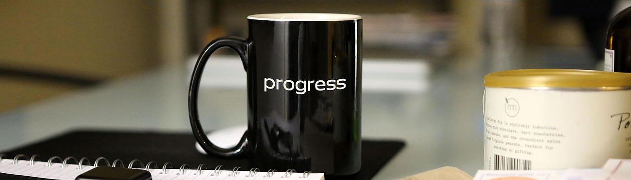 Tasse mit Aufdruck "progress" auf einem Schreibtisch.
